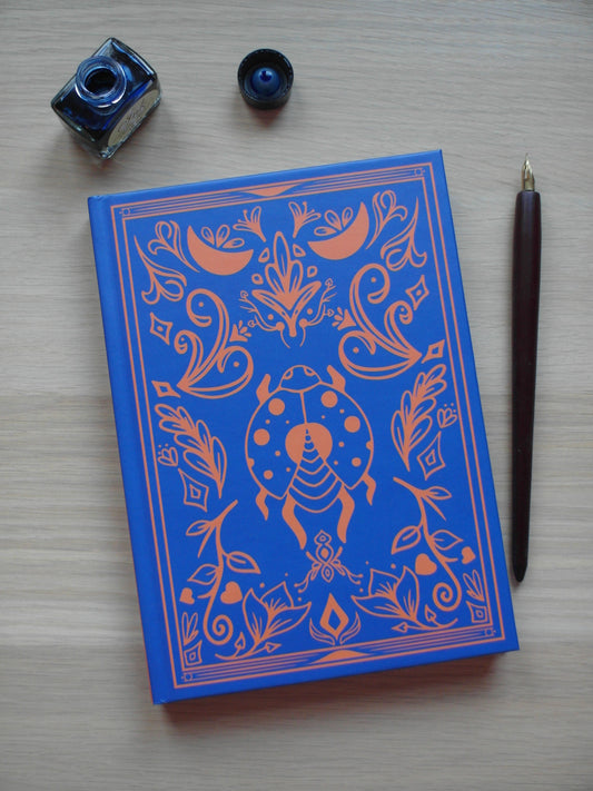 blue and orange hardback journal featuring designs of floral flourishes and swirls around a central beetle - image decor of a pen and ink pot